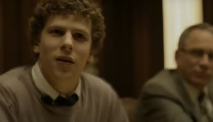 Young man with curly hair and a man in glasses in a dimly lit room, scene from the greatest films of the new millennium.