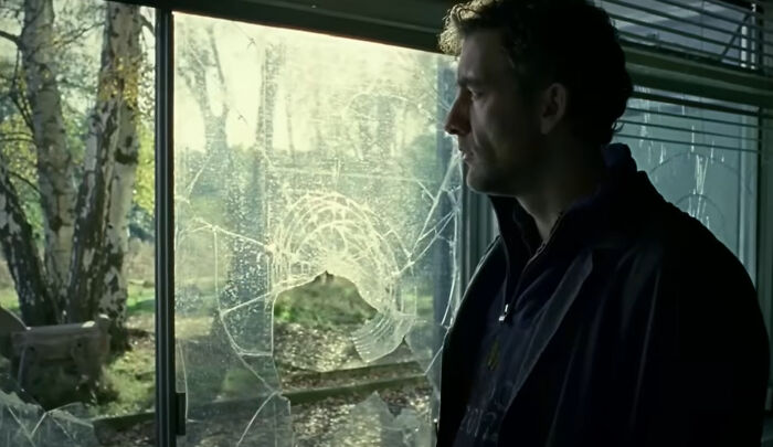Man in dark jacket looks through shattered window in a dramatic scene from one of the new millennium's greatest films.