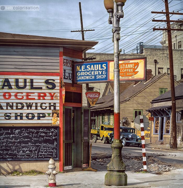 Colorized historical photo of an old grocery and sandwich shop on a street corner with vintage cars nearby.