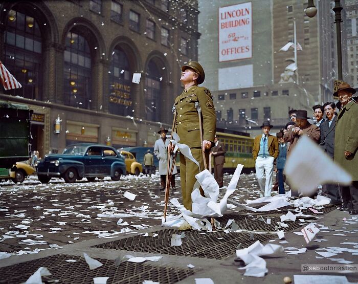 Colorized historical photo of a soldier on crutches surrounded by paper debris in a busy urban street scene.