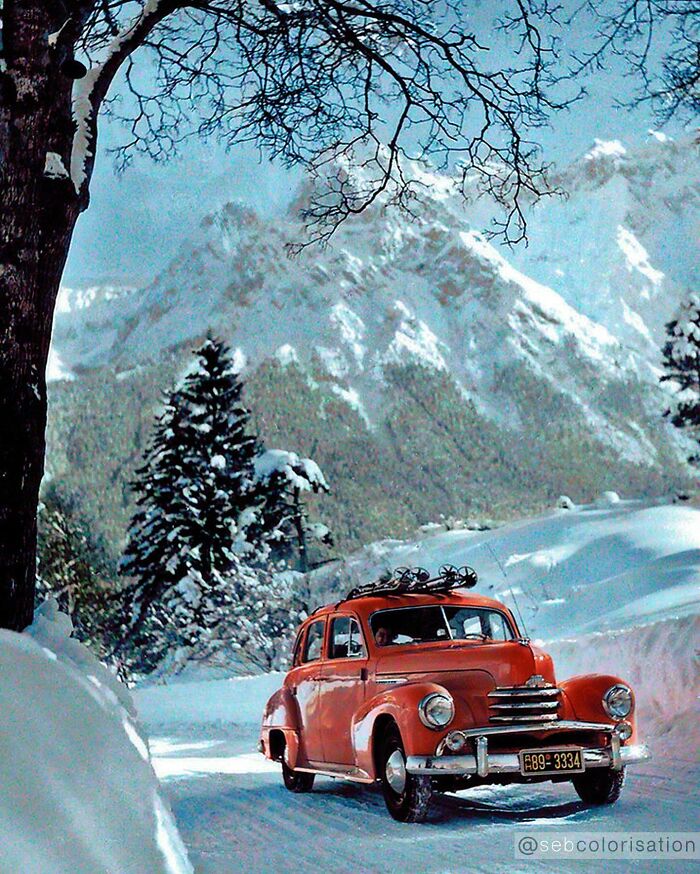 Vintage red car driving on snowy mountain road with snow-covered trees, colorized historical photo by talented viral artist