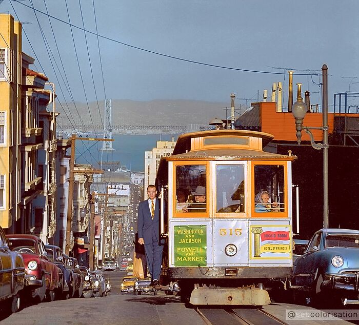 Colorized historical photo of a San Francisco cable car on a street with passengers and vintage cars in the background.