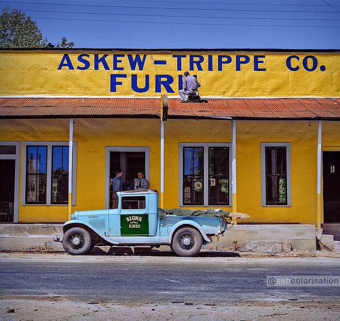Colorized historical photo of a worker painting a sign on a bright yellow building with a vintage blue truck parked outside.