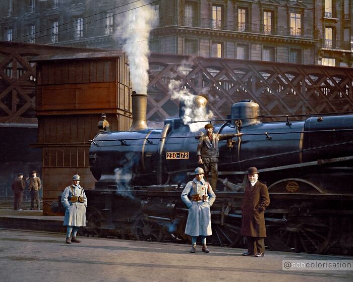 Colorized historical photo showing workers and officers near a steam locomotive at an urban train station platform.