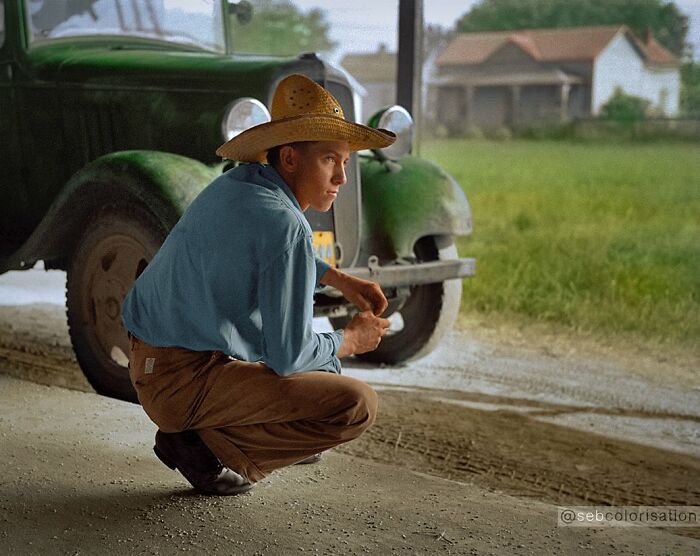 Young man in a straw hat crouching by a vintage green truck in a rural setting, colorized historical photo.