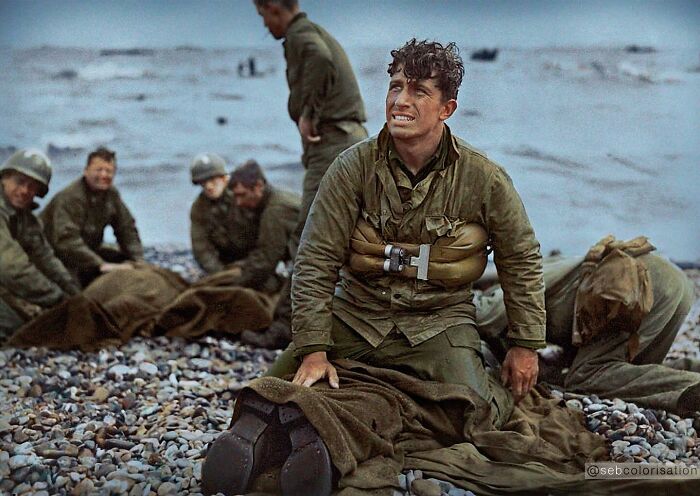Colorized historical photo showing a group of soldiers on a rocky beach during wartime, with emotional expressions.
