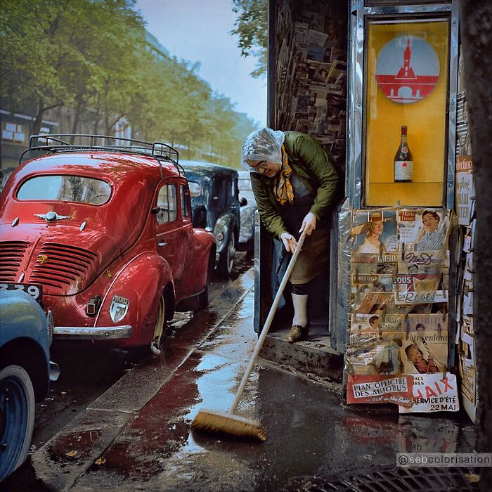 Woman sweeping outside a shop on a rainy street, vintage cars parked, colorized historical photo by viral artist.