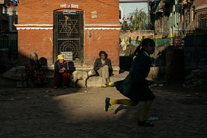 Candid street photo of a girl running past seated people and a dog, capturing the quiet beauty of everyday life outdoors.