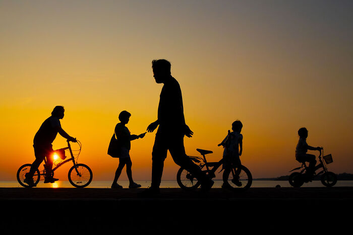 Silhouettes of people and children on bikes at sunset, candid street photo capturing the quiet beauty of everyday life.