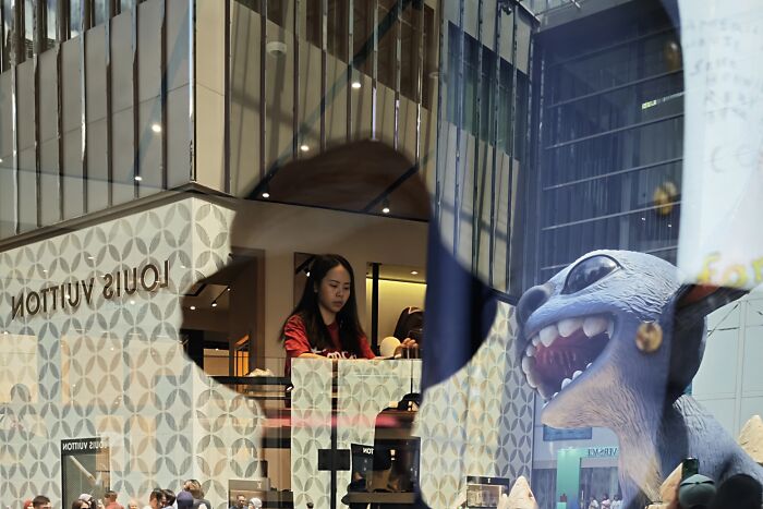 Candid street photo capturing quiet beauty with a young woman inside a luxury store and reflections of urban life outside.