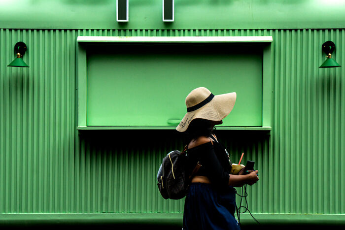 Person wearing a wide-brimmed hat walking past a green wall, capturing candid street photos of everyday life moments.