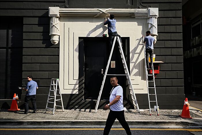 Three workers on ladders fixing a building facade and a man walking by in a candid street photo capturing everyday life.