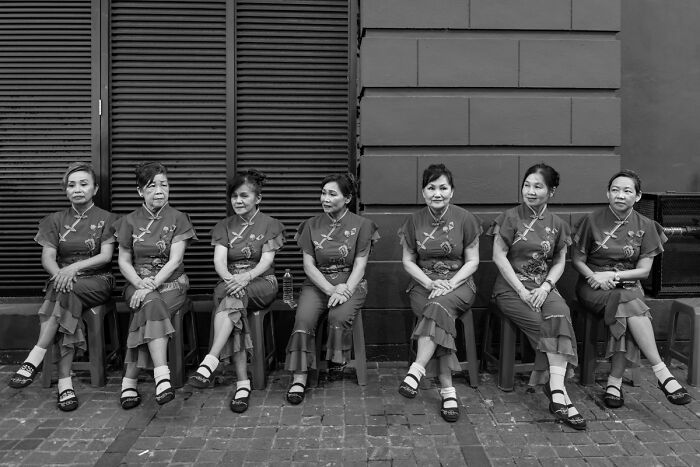 Seven women in traditional dresses sitting quietly on chairs by a wall in candid street photos capturing everyday life.