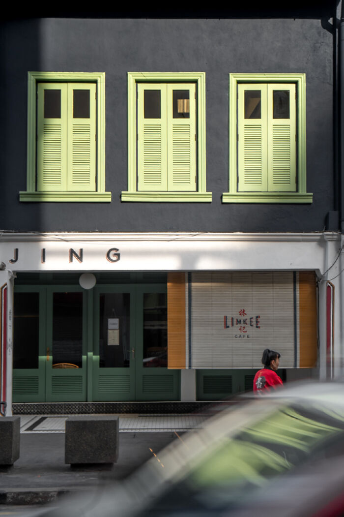 Candid street photo of a person walking past a building with green shutters, capturing quiet beauty of everyday life.