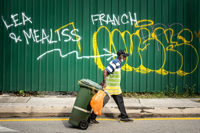 Man wearing a mask pulling a green trash bin on a street with colorful graffiti, candid street photo of everyday life.