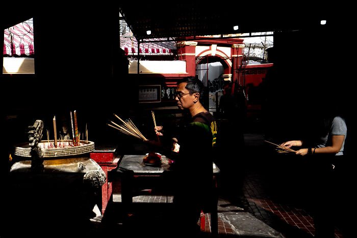 Man lighting incense sticks inside temple in candid street photo capturing quiet beauty of everyday life in urban setting.