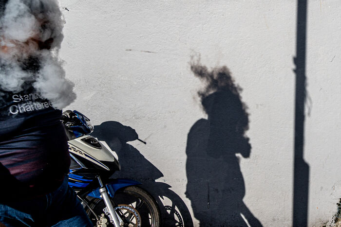 Candid street photo of a person on a motorcycle with smoke and dramatic shadows on a white wall.