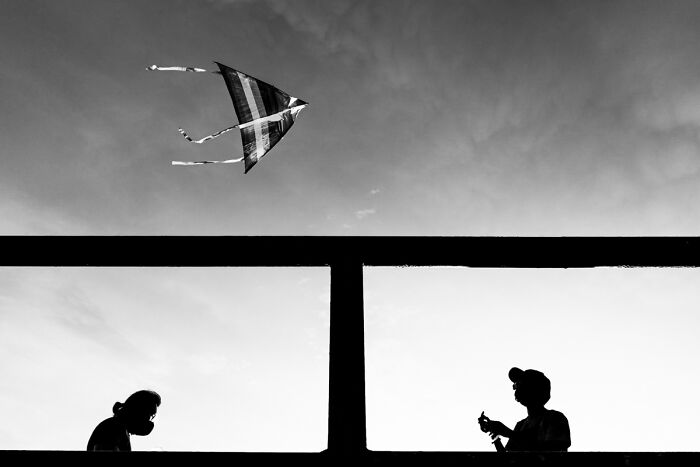 Silhouettes of two people on a bridge with a kite flying above, candid street photo capturing quiet everyday life.