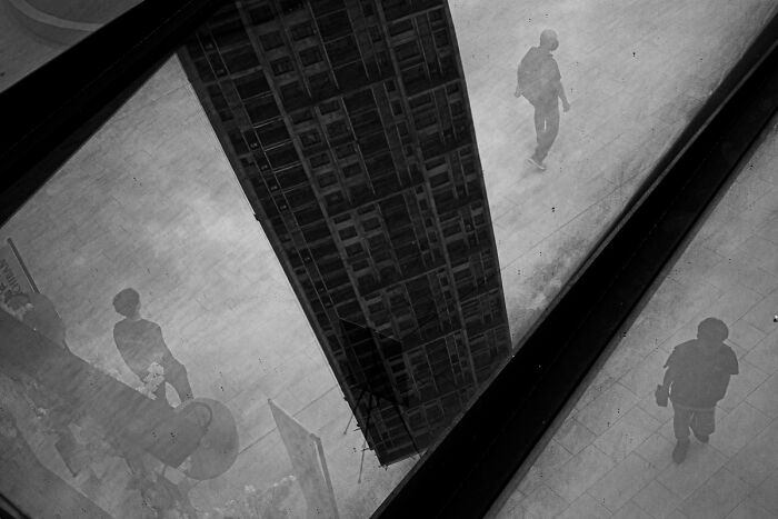 Candid street photo showing reflections of pedestrians and a building, capturing the quiet beauty of everyday life.