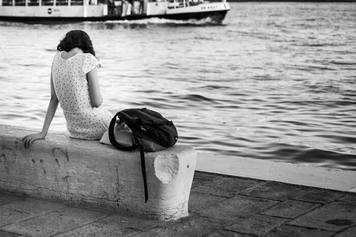Young woman in a patterned dress sitting by the water with a backpack, captured in a cinematic photo by Adriana Ferrarese.
