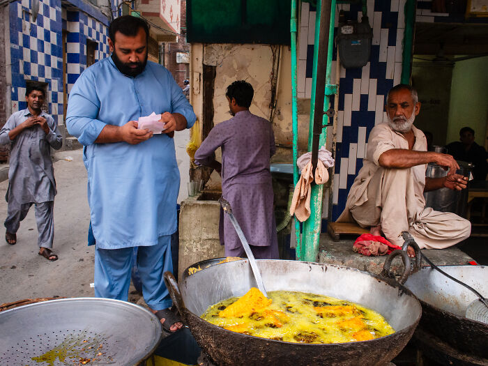 Man in blue traditional clothes standing near a street food vendor frying snacks in a busy candid street moment full of emotion.