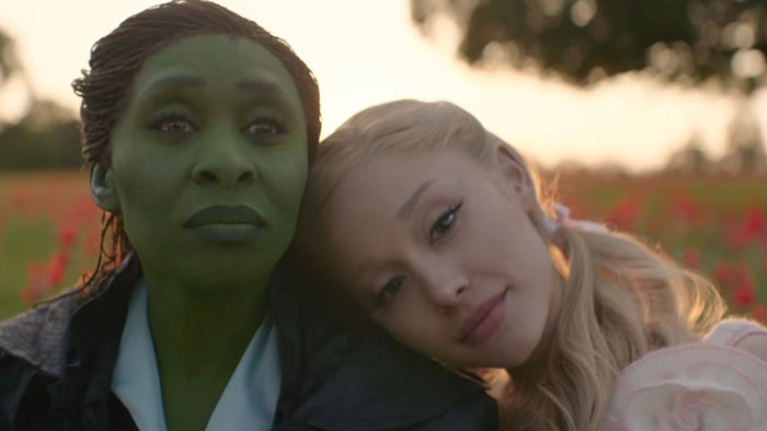 Two main characters from the new Wicked movie, one with green makeup and another as Dorothy, in an outdoor scene.