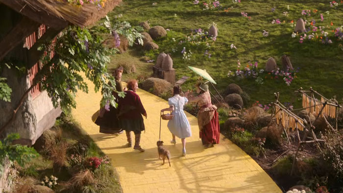 Scene from Wicked movie showing characters including Dorothy walking on the yellow brick road in a fantasy setting.