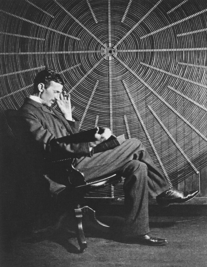 Nikola Tesla seated and reading in front of a large circular coil, representing his impactful contributions to science.