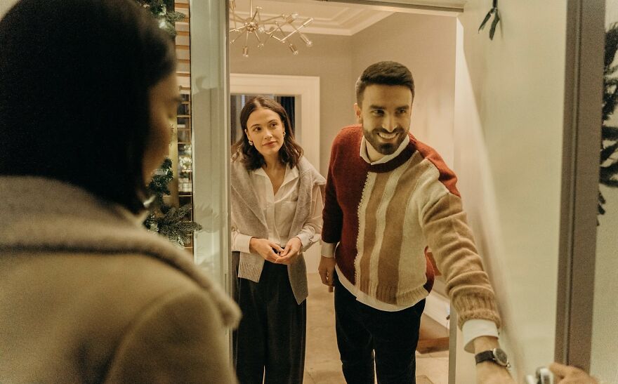 Couple welcoming a guest at the door indoors, smiling and engaging in a friendly then vs now poll conversation.