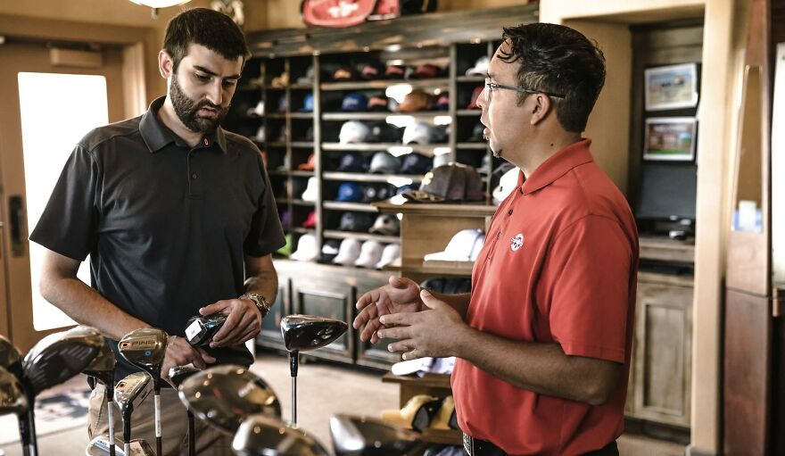 Two men discussing golf equipment at a store counter in a casual setting for then vs now poll.
