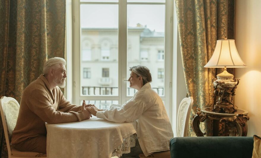 Elderly couple holding hands across table in cozy room, reflecting on moments for then vs now poll discussion.