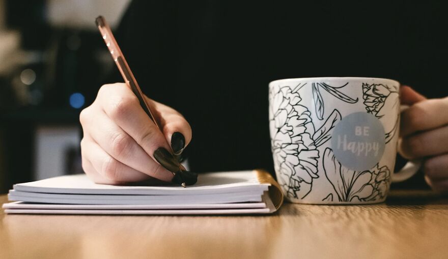 Person holding a pen and writing in a notebook next to a floral mug with a Be Happy message for then vs now poll.