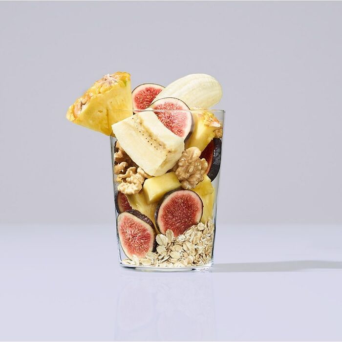 Glass filled with fresh figs, banana, pineapple, walnuts, and oats, a mouthwatering food photo by Kai Stiepel.