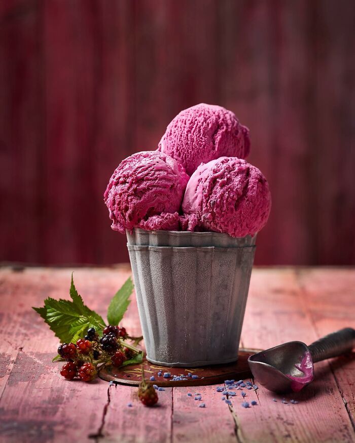 Three scoops of vibrant pink ice cream in a metal cup with fresh berries on a rustic wooden surface, food photography.