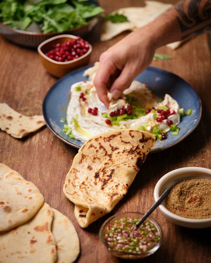 Hand dipping naan bread into creamy dip with pomegranate seeds and herbs in a mouthwatering food photo by Kai Stiepel.