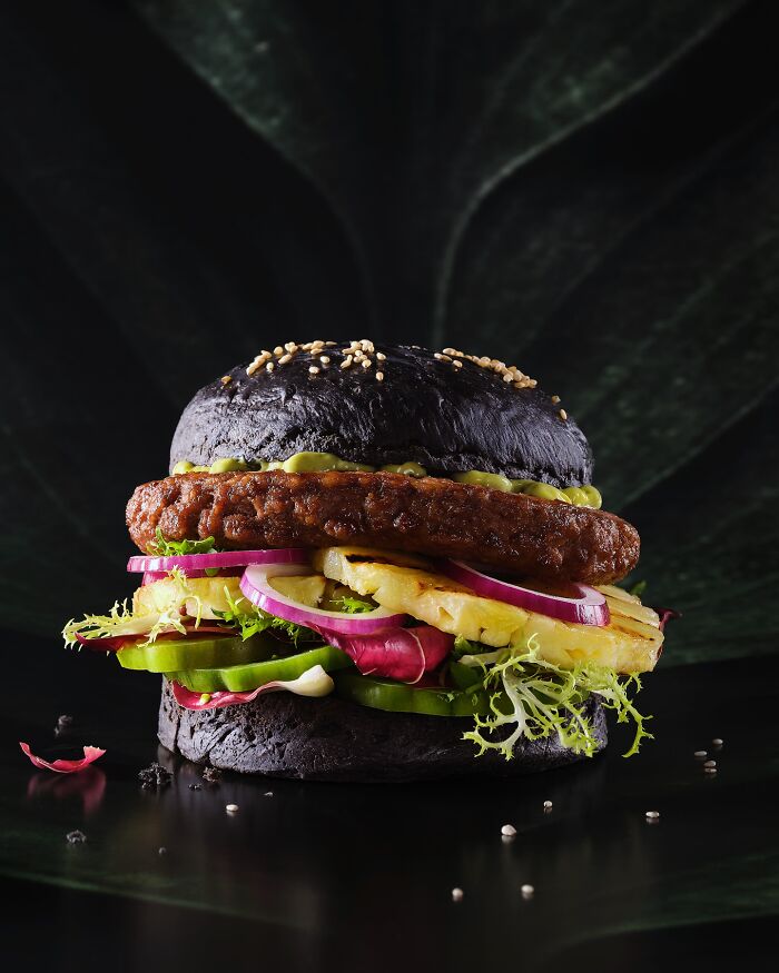 Black bun burger with grilled pineapple, fresh vegetables, and sauce, a mouthwatering food photo by Kai Stiepel.