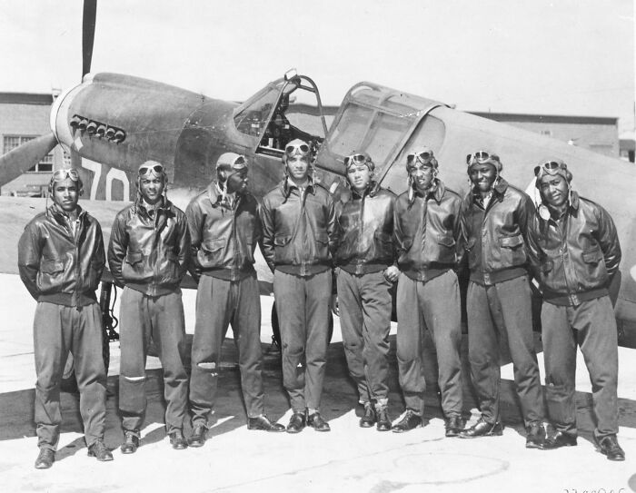 Group of African American military pilots in leather jackets and goggles standing in front of a vintage aircraft in military history context