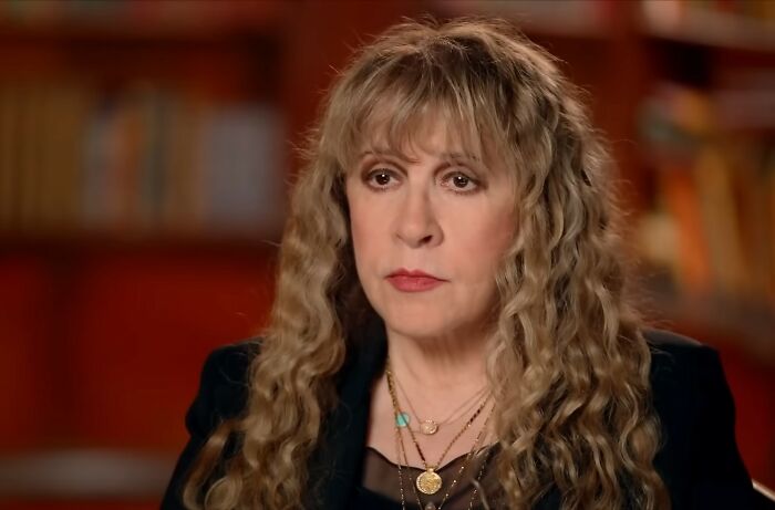 Woman with long curly hair wearing layered necklaces and dark outfit, speaking about pregnancy and twins in an indoor setting.