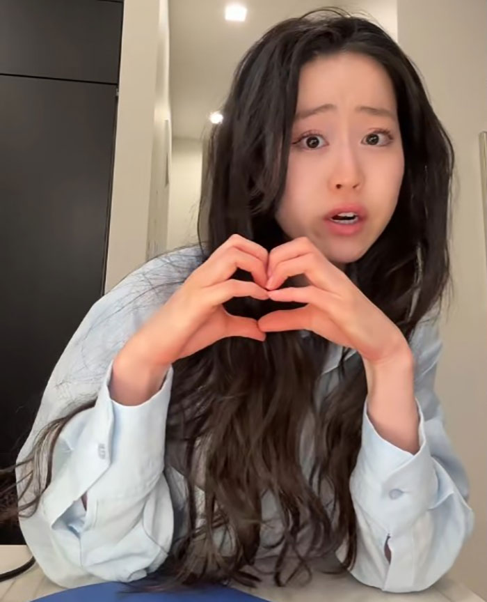 Young woman with long dark hair making a heart shape with her hands, reacting to Diddy's aged look in court.