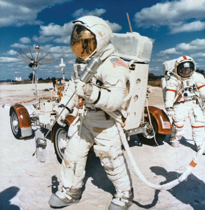 Astronauts in full space suits testing lunar rover on a sandy terrain under a blue sky with clouds.