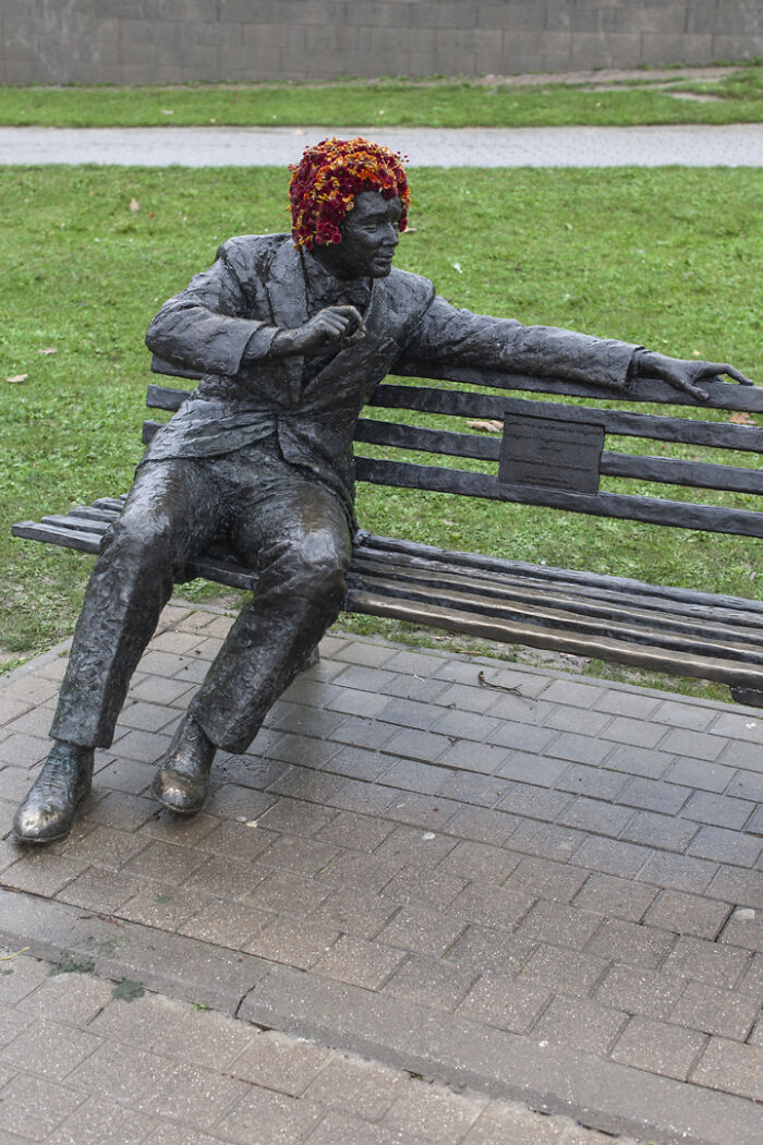 Bronze city statue dressed in flowers with floral hair sitting on a bench in an outdoor park setting.