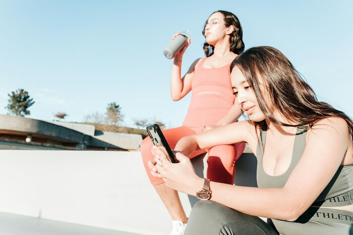 Two women in activewear outdoors, one checking her phone, illustrating behaviors that may indicate someone is insecure.