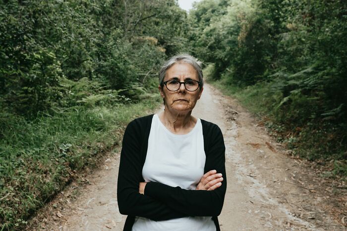 Older woman over 42 standing on a forest path with arms crossed, reflecting strength and resilience.