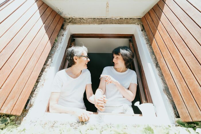 Two women over 42 wearing white shirts, smiling and talking while leaning out of a wooden window frame.