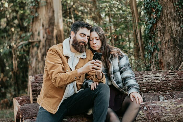 A couple sitting on logs in the forest, looking at a smartphone, discussing twins pregnancy facts and experiences.