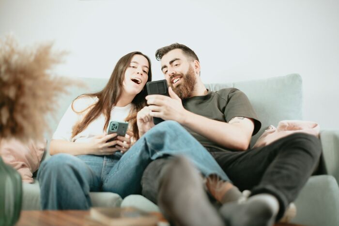 Young man and woman sitting on a couch smiling and using smartphones illustrating behaviors that may indicate insecurity.