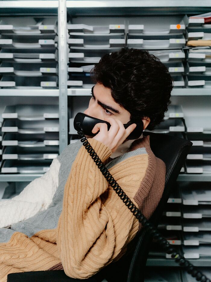 Young man using a landline phone, reflecting on childhood joys and life before smartphones in a busy office setting.