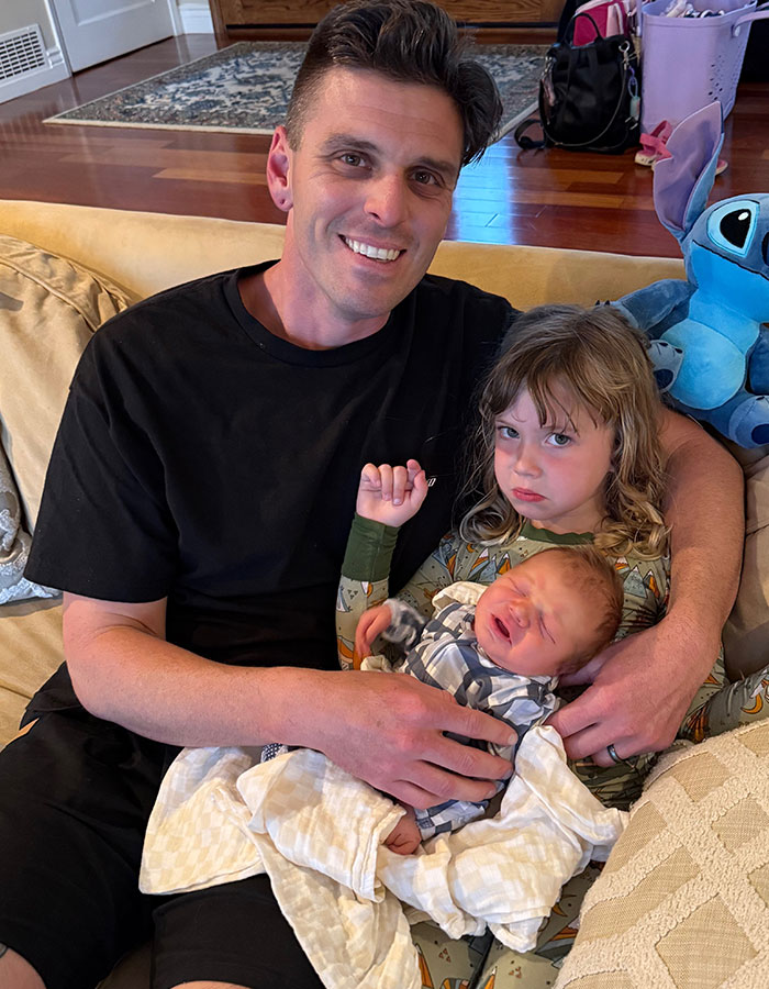 Man sitting on a couch holding two children, capturing a tender family moment after a real housewives exec childbirth loss.