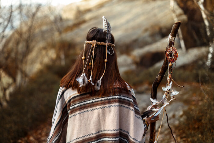 Person wearing feathers and beads in hair, holding a decorated walking stick, reflecting on unusual baby names outdoors.