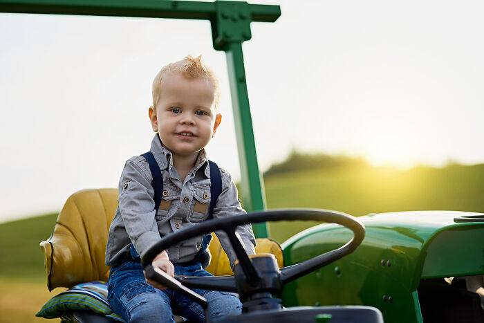 Toddler boy with suspenders sitting on green tractor outdoors, illustrating unique baby names that secretly weird people out.